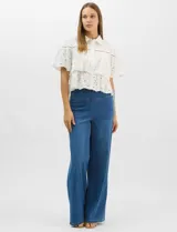 Camisa de algodón blanca con cuello clásico y cierre frontal de botones. Presenta un diseño con capas superpuestas y volados, combinando paneles de tela lisa con tejido bordado suizo de motivos calados y una cenefa decorativa. Posee mangas cortas y una silueta acampanada.