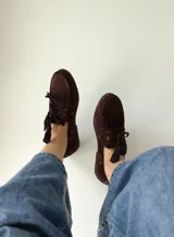 Mocasín tipo náutico de gamuza color marrón oscuro, con cordones frontales y detalle de flecos decorativos en el empeine.