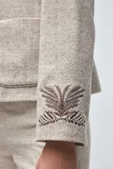 Chaqueta de lana fina sin teñir, con cuello clásico y bolsillos frontales. Presenta detalles bordados en el cuello y en los puños de las mangas. Es una prenda forrada con diseño exclusivo.