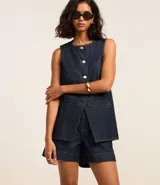 Chaleco de jeans azul oscuro, sin mangas, con cuello redondo y cierre frontal de botones blancos. Presenta un corte ligeramente alargado y solapas frontales que simulan bolsillos.