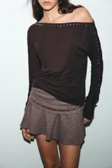 Camiseta de cuello barco y manga larga tipo murciélago, confeccionada en tejido de punto gris melange. Presenta detalle de tachas metálicas a lo largo de los hombros y las mangas.