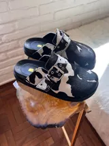 Mules con estampa de vaca en blanco y negro, con capellada cerrada, tira ajustable con hebilla metálica y ojales decorativos.