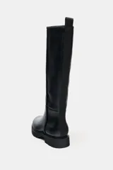 Bota de caña alta estilo plano, confeccionada en material sintético negro con acabado liso. Presenta detalle de pespuntes verticales en el cuerpo, punta redonda, suela track dentada y tirador trasero para facilitar el calce.