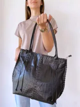 Cartera tipo tote de cuero negro con textura de cocodrilo. Presenta detalles de costuras trenzadas a lo largo de la estructura y tiras decorativas laterales. Cuenta con doble asa de hombro y cierre superior.