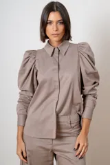 Camisa de algodón color topo, con cuello clásico, cierre frontal oculto y mangas largas abullonadas con puños ajustados. Presenta botones decorativos con strass en el cuello y los puños.