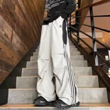 Pantalón deportivo color beige claro, estilo baggy o ancho, con cordón ajustable en la cintura y cierre completo impermeable (full zip) en los laterales. Presenta una franja vertical negra con tres líneas blancas a lo largo de la costura lateral, y un pequeño logo bordado en la parte superior del muslo.