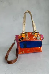 Cartera de cuero de diseño exclusivo con intervención artística de Carolina De Cunto, combinando tonos naranja, azul y detalles en color natu y terra. Cuenta con doble asa de hombro, solapa frontal de pelo sintético azul y herrajes metálicos.