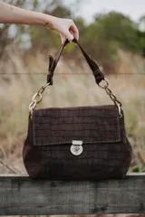 Cartera de mano con solapa, confeccionada en cuero con textura tipo cocodrilo. Presenta una manija corta con eslabones metálicos decorativos y cierre frontal con herraje metálico a presión.