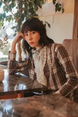 Camisa de manga larga con cuello alto y pechera adornada con volantes de tul bordado. El tejido principal es de algodón con estampado a cuadros en tonos marrón, blanco y azul claro.