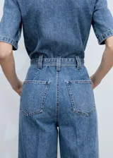 Mono largo de denim azul con cuello camisero, mangas cortas, cierre frontal con cremallera oculta, cinturón ajustable con hebilla metálica, bolsillos laterales y un bolsillo de parche en el pecho.