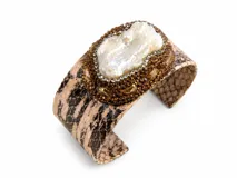 Pulsera tipo brazalete rígido con textura de piel de reptil en tonos beige y marrón. Presenta un aplique central destacado compuesto por una perla barroca rodeada de cristales y strass en tonos marrones y transparentes.