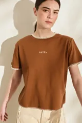 Remera marrón de manga corta y cuello redondo, con detalles de costuras decorativas en contraste color crudo en el cuello, los puños y el dobladillo inferior. Cuenta con el nombre de la marca impreso en el pecho.