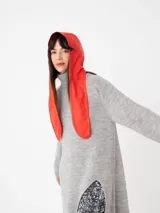 Buzo largo gris de tejido de punto grueso con cuello alto y capucha con orejas de conejo rojas. Tiene mangas largas y un tajo lateral.