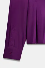 Camisa de manga larga en color violeta, con cuello solapa y cierre frontal de botones. Presenta un diseño de corte recto con detalle de pliegues en la espalda.
