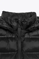 Chaleco acolchado de color negro con cuello subido, capucha ajustable y cierre frontal con cremallera termosellada. Presenta un diseño con paneles laterales en tejido liso y bolsillos laterales.