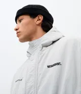 Campera bomber de poliamida en color gris claro, con capucha, cierre frontal con botones a presión y lettering bordado en el pecho. Presenta un diseño funcional con detalles utilitarios.