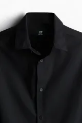 Camisa de vestir negra con cuello inglés, tapeta clásica y mangas largas con puños ajustables. Presenta un corte entallado que se ajusta al cuerpo y bajo levemente redondeado.