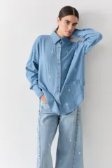 Camisa de jean azul claro con bordados pequeños en color blanco de figuras deportivas. Presenta cuello clásico, cierre frontal con botones de madera y mangas largas con puños abotonados.