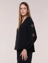 Blusa holgada de color negro, confeccionada en 100% poliéster con textura liviana. Cuenta con escote en V, mangas largas raglán con detalles de calados en forma de flores y puños elastizados.