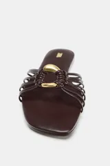 Sandalia plana de mujer, color marrón oscuro, con puntera cuadrada. Presenta un diseño de tiras finas que se unen en el centro con un adorno anillado metálico dorado.