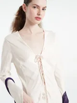Vestido largo tipo kaftan color crema, confeccionado en una mezcla de cupro y rayón. Presenta un escote en V profundo con detalle de cordones cruzados y mangas largas adornadas con flecos largos en color violeta.