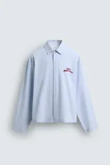 Camisa de algodón de corte regular, color celeste con finas rayas blancas verticales. Presenta un bordado en color rojo en el pecho con la frase "Café du Matin Doux".