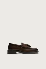 Mocasín de vestir color marrón oscuro, confeccionado en cuero, con detalle de antifaz y borlas decorativas en el empeine. Presenta una horma redonda y suela plana a tono.