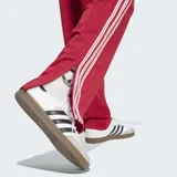 Pantalón deportivo Adidas Firebird Drop Needle de color rojo con rayas verticales finas, bolsillos laterales con cierre y las icónicas tres tiras blancas a lo largo de los laterales. Incluye el logo del trifolio bordado en la pierna.