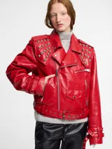 Chaqueta biker roja de cuero sintético con tachas plateadas en cuello y hombros, cierre asimétrico con cremallera, cinturón con hebilla y múltiples bolsillos con cremallera.