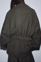 Chaqueta fluida de color verde oliva, con cuello solapa, manga larga ajustable con trabilla y botón, y cinturón del mismo tejido anudado a la cintura.