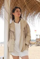 Chaqueta de gamuza color beige con diseño de flecos en el frente y las mangas. Presenta solapas clásicas, cierre frontal con botones y bolsillos laterales.