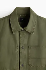 Sobrecamisa de lona de algodón en color verde militar, con cuello clásico, cierre frontal de botones y tres bolsillos de parche frontales. Presenta mangas largas con puños abotonados y un diseño de corte recto.