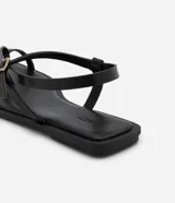 Sandalias planas de cuero sintético negro, con puntera cuadrada, tira delantera con dedera y tira trasera con hebilla metálica dorada.