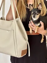Bolso tipo tote de cuero color beige con doble asa de hombro y bolsillo externo con botón metálico.
