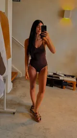 Traje de baño entero color marrón oscuro, con escote en V y tirantes finos que se cruzan en la espalda. Presenta detalles de ajuste o decoración en los laterales de la cadera.