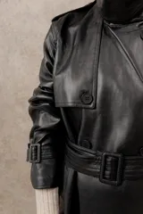 Trench largo de ecocuero negro, con cuello clásico, cierre cruzado con botones laterales y cinturón ajustable.
