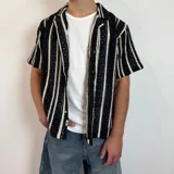 Camisa de crochet a rayas verticales blancas y negras, con cuello clásico, corte boxy y mangas cortas.