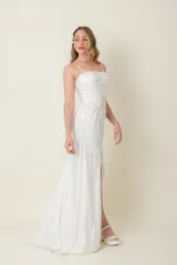 Vestido largo blanco de satén de seda natural, con breteles finos, escote recto, drapeado en el busto, tajo lateral y cola corta. Presenta aplique de flor de organza bordada en un costado.