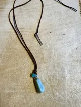 Collar largo con tiras de cuero sintético tostado y dije de piedra en bruto de tonalidad rojiza con detalles metálicos, acompañado de una placa metálica grabada.