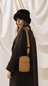 Bandolera pequeña de diseño compacto en color mostaza, confeccionada en material sintético con textura granulada. Presenta un bolsillo frontal con el logo de la marca impreso, cierre superior y correa ancha ajustable y desmontable.