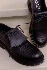 Mocasín de cuero vacuno negro con textura tipo reptil, suela track de goma, cordones finos y solapa con flecos sobre el empeine.