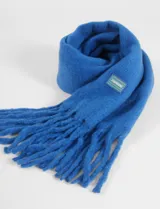 Bufanda de textura suave y acabado afelpado, color negro, con flecos en los extremos y un pequeño parche cuadrado decorativo con logo en color verde y azul.