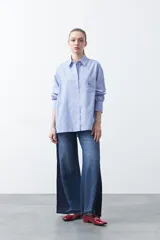 Camisa a rayas azules y rojas de manga larga, confeccionada en tejido yarn dye. Presenta cuello camisero, botones frontales y un bordado distintivo en el bolsillo del pecho.