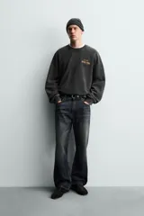 Sudadera de corte relajado, color negro con efecto lavado, cuello redondo y manga larga. Presenta un pequeño estampado frontal con texto en tonos naranja y amarillo.