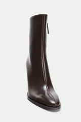 Botín de cuero color marrón oscuro, con tacón fino alto, punta fina y cierre lateral. Presenta un diseño minimalista con costuras visibles en el empeine.