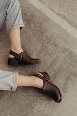 Zuecos de cuero color marrón oscuro, con diseño cerrado en la parte delantera, tira ajustable con hebilla metálica en el talón y taco cuadrado bajo.
