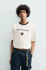 Camiseta de corte relajado confeccionada en algodón, color blanco crudo con cuello redondo en contraste color negro. Presenta un bordado frontal con la frase 'Coração de Melão' y un parche en forma de corazón.