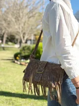 Mini bandolera de cuero con diseño de flecos largos y hebilla frontal decorativa. Presenta un acabado texturizado con efecto craquelado en tonos blanco y marrón, y correa ajustable.