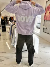 Canguro oversize lila con capucha y gran estampado blanco en la espalda con la palabra "LOVE" y un imperdible.