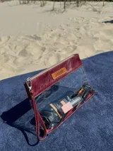 Necessaire transparente con detalles en cuero bordó, cierre superior con cremallera y correa de mano.